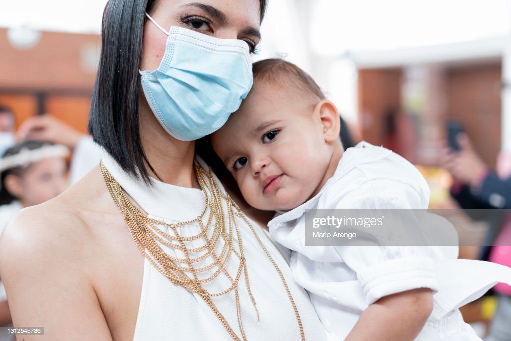 mamá latina con máscara lleva a su bebé de 1 año tanto en la iglesia donde se celebra el bautizo del bebé - bautizo fotografías e imágenes de stock