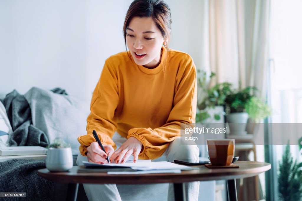 young asian woman holding a pen and signing paperwork in the living room at home. deal concept - escribir fotografías e imágenes de stock