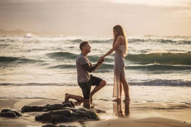 joven proponiendo a su novia en una playa - compromiso fotografías e imágenes de stock
